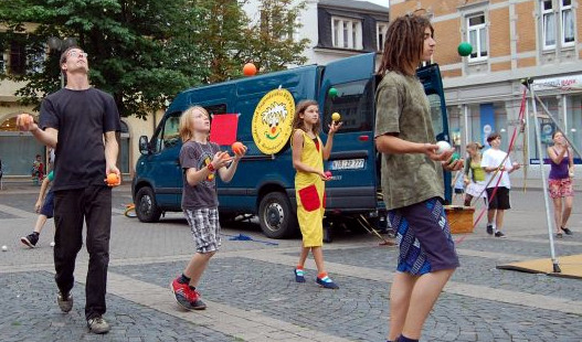 Zirkus Pepperoni Jonglage & Artistik zum Mitmachen - Kinder mit unterschiedlichem Alter jonglieren mit bunten Bällen. Dahinter steht ein blauer Bus vom Zirkus.