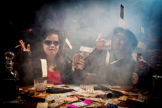 Eine Frau und ein Mann mit dunklen Sonnenbrillen sitzen an einem Pokertisch. Sie werfen Geldscheine in die Luft.