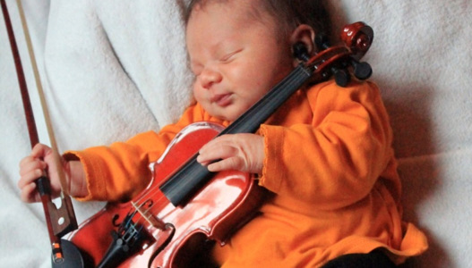Ein Baby liegt auf einer kuscheligen Decke. Es hat die Augen geschlossen. In den Händen hält es eine Geige und einen Geigenbogen.