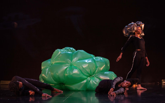 Panaibra Gabriel Canda Metamorphoses, eine Tänzerin mit einer Kopfbedeckung aus goldenen Plastiksäcken schreitet über die Bühne, im Hintergrund eine grüne Plastikskulptur