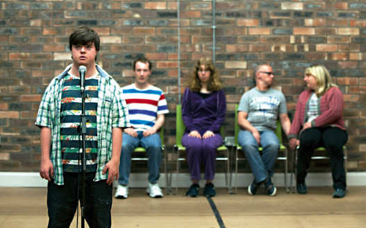 Mind the Gap Contained, vier Darsteller sitzen vor einer Mauer, ein weiterer steht im Vordergrund an einem Mikrofon