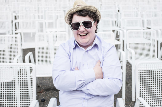 Musiker Carsten Schnathorst sitzt in einem leeren Stadion alleine auf einem weißen Sitz. Er hat einen Hut auf und eine große dunkle Sonnenbrille.