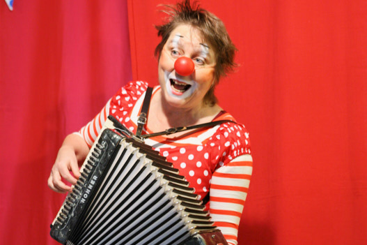Clownin Rosina steht vor einem roten Vorhang. Sie hat eine Clownsnase auf und spielt auf einem Akkordeon, dabei singt sie.