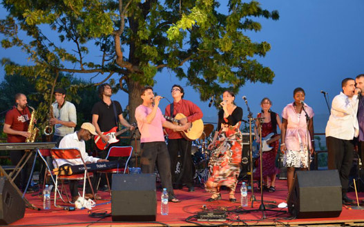 Percujam - Elf Musiker und Musikerinnen stehen auf einer Bühne unter einem großen Baum. Sie singen in Mikrofone, manche von ihnen spielen Instrumente