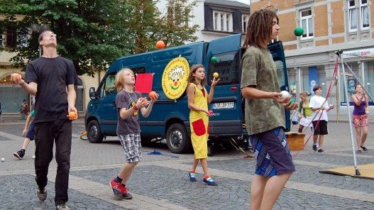 Zirkus Pepperoni Jonglage & Artistik zum Mitmachen - Kinder mit unterschiedlichem Alter jonglieren mit bunten Bällen. Dahinter steht ein blauer Bus vom Zirkus.
