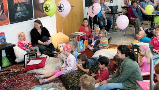 Viele Kinder und ihre Eltern sitzen auf Kissen im Foyer des Staatstheaters auf dem Boden und hören einer Vorleserin zu